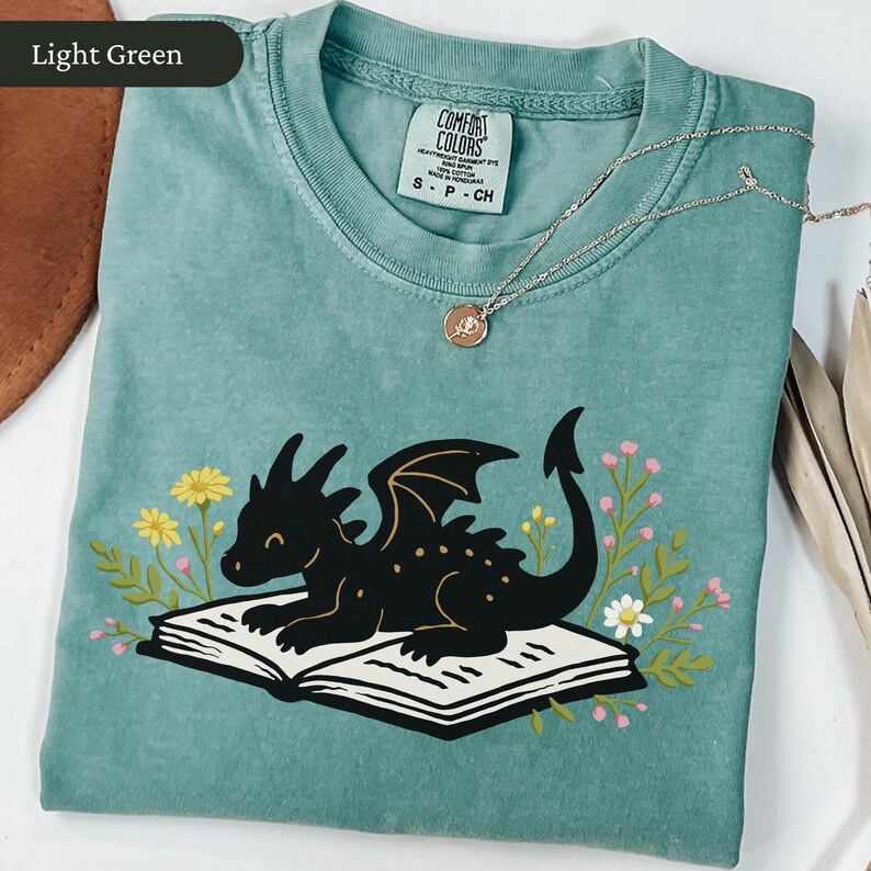 Puede incluir: Camiseta verde claro con una ilustraci&oacute;n de un drag&oacute;n negro tumbado sobre un libro abierto, rodeado de flores. La camiseta tiene cuello redondo y mangas cortas. Un collar plateado con un peque&ntilde;o colgante est&aacute; sobre la camiseta.