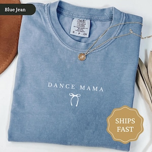 May include: A blue jean coloured t-shirt with the words "DANCE MAMA" in white script. A gold necklace with a pendant rests on the shirt. The shirt has a crew neck and short sleeves. The shirt is made of a soft, comfortable material.