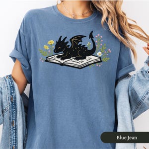 Puede incluir: Camiseta azul vaquero con una ilustraci&oacute;n de un drag&oacute;n negro sobre un libro abierto, rodeado de flores. El drag&oacute;n tiene alas y sonr&iacute;e. La camiseta tiene las mangas enrolladas.