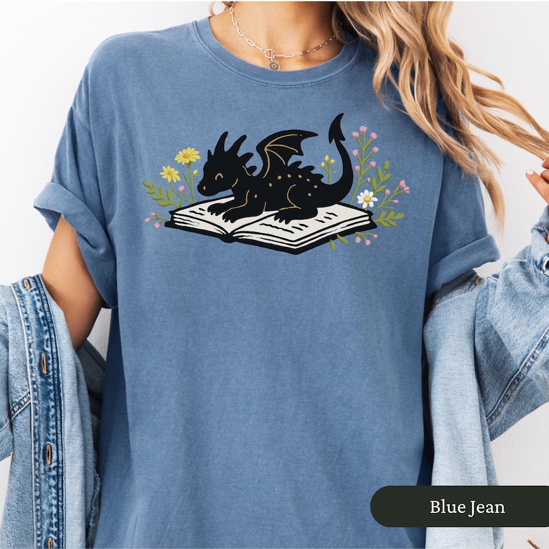 Puede incluir: Camiseta azul vaquero con una ilustraci&oacute;n de un drag&oacute;n negro sobre un libro abierto, rodeado de flores. El drag&oacute;n tiene alas y sonr&iacute;e. La camiseta tiene las mangas enrolladas.