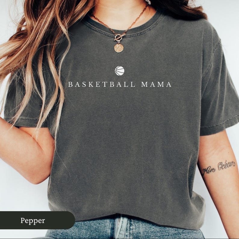 以下が含まれることがあります： 「BASKETBALL MAMA」の文字とバスケットボールのグラフィックが入った濃いグレーのTシャツ。クルーネックのTシャツで、柔らかく快適な素材で作られているようです。Tシャツは無地です。