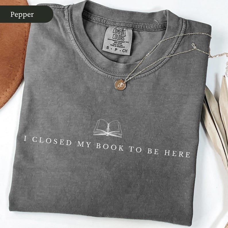 May include: Pepper-colored t-shirt with the text "I CLOSED MY BOOK TO BE HERE" and a book graphic. A silver necklace with a pendant is draped across the shirt. The shirt has a Comfort Colors tag.