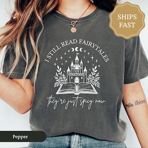 May include: Dark grey t-shirt with a white graphic. The design includes a castle on an open book, stars, moons, and the text "I STILL READ FAIRYTALES" and "they're just spicy now."