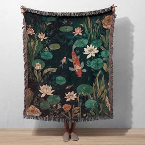 K&ouml;nnte beinhalten: Eine gewebte Wurmdecke mit einer dunkelgr&uuml;nen Teichszene mit Koi-Karpfen, Seerosen und Lotusblumen. Die Decke hat einen Fransenrand mit Braun- und Beiget&ouml;nen. Die Koi-Karpfen sind orange und wei&szlig;.