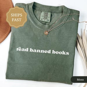 May include: A moss green t-shirt with the words "read banned books" printed in white. The shirt has a crew neck and short sleeves. A gold necklace with a small pendant rests on the shirt. The shirt is made of a soft, comfortable material.