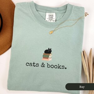 Może przedstawiać: Jasnozielony t-shirt z czarnym kotem siedzącym na stosie książek. Na koszulce znajduje się napis "cats & books."