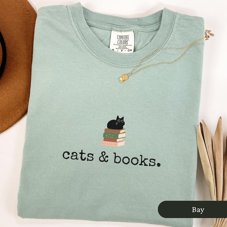 Może przedstawiać: Jasnozielony t-shirt z czarnym kotem siedzącym na stosie książek. Na koszulce znajduje się napis "cats & books."