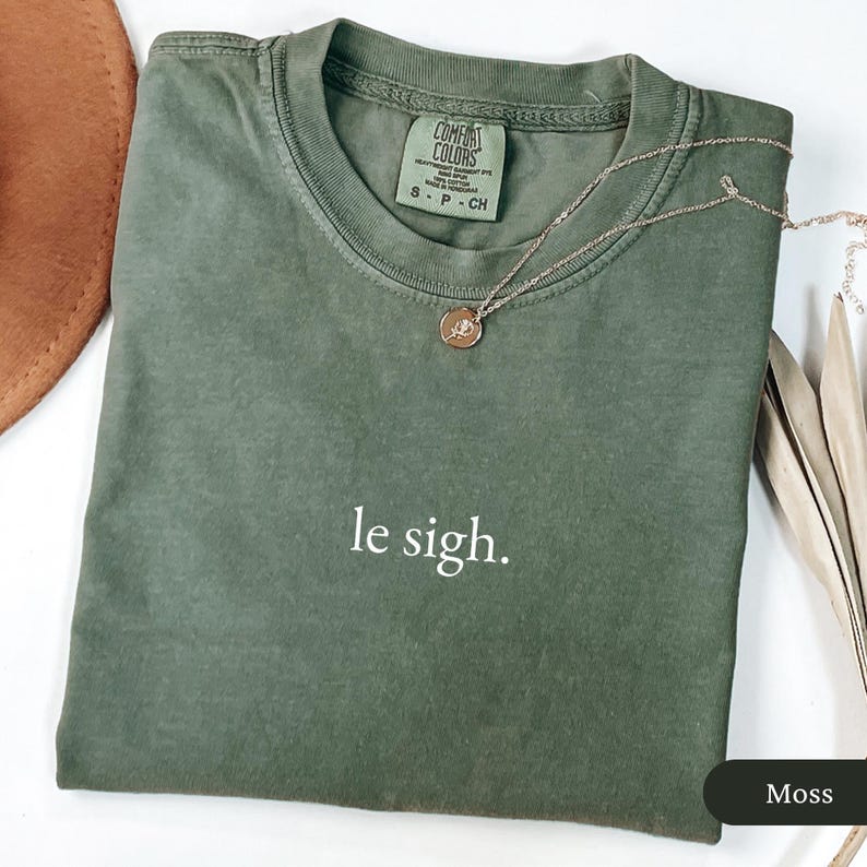 Pu&ograve; includere: Una maglietta verde muschio con la frase "le sigh" stampata in bianco. Una collana color oro con un piccolo ciondolo &egrave; appoggiata sulla maglietta. La maglietta &egrave; piegata e ha un'etichetta con scritto "Comfort Colors".