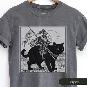 May include: A gray t-shirt with a black and white illustration of a knight riding a black cat. The knight is wearing armor and holding a sword. The cat has a collar and is looking to the right. The illustration is in a square frame and the text "Pepper" is at the bottom of the shirt.