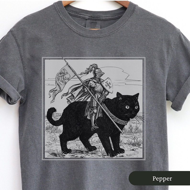 May include: A gray t-shirt with a black and white illustration of a knight riding a black cat. The knight is wearing armor and holding a sword. The cat has a collar and is looking to the right. The illustration is in a square frame and the text "Pepper" is at the bottom of the shirt.
