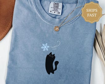 May include: Blue jean colored Comfort Colors t-shirt with a black cat graphic and a snowflake. The shirt has a crew neck and short sleeves. A gold necklace with a pendant is draped across the shirt.