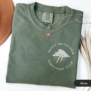 May include: Moss green t-shirt with a crew neck and short sleeves. The shirt features a graphic of a cloud with lightning bolts and the text "DAILY SHITSTORM SURVIVORS CLUB". A gold necklace with a pendant is on the shirt.