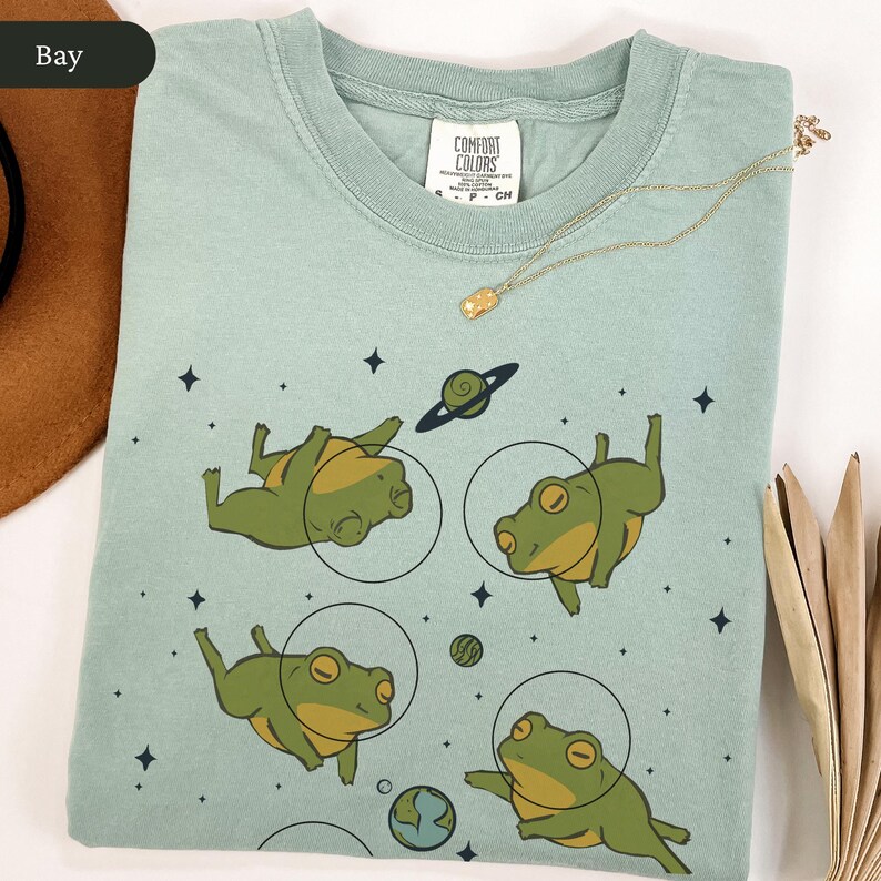 May include: A light green Comfort Colors t-shirt featuring a whimsical space-themed design. The shirt showcases four cartoon frogs floating in space, each within a circular frame, alongside planets and stars. A gold necklace rests on the shirt.