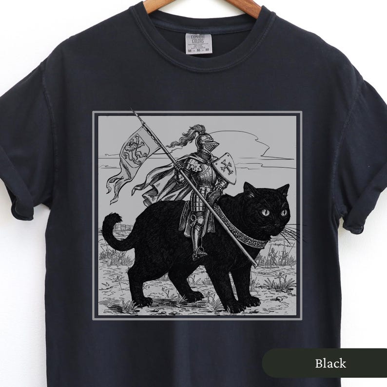 May include: Black t-shirt with a graphic print of a knight riding a black cat. The knight is holding a sword and a flag with a lion on it. The text "Black" is printed on the bottom right corner of the shirt.