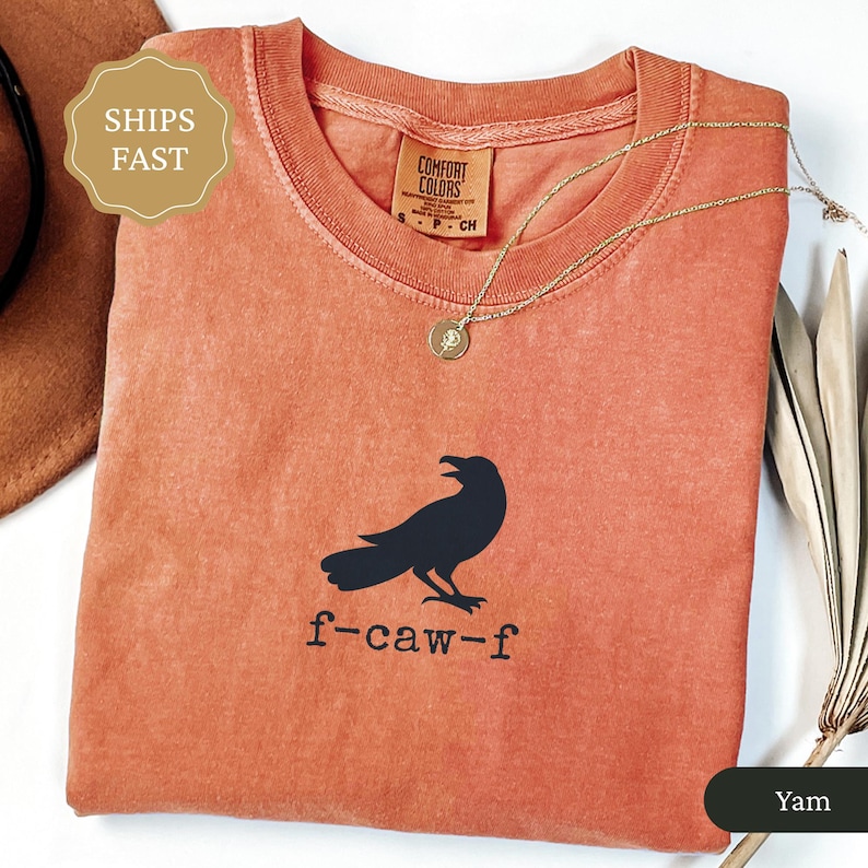 May include: A coral-colored t-shirt with a black crow graphic and the text "f-caw-f". The shirt has a crew neck and short sleeves. A gold necklace with a pendant is draped on the shirt.