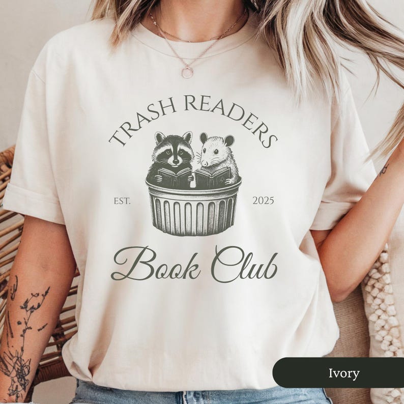 Puede incluir: Una camiseta blanca con el texto "Trash Readers Book Club" en negro. El texto est&aacute; rodeado de un c&iacute;rculo con las palabras "Est. 2025". Dentro del c&iacute;rculo hay ilustraciones de un mapache y una rata leyendo libros en un contenedor de basura.