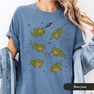 May include: Blue jean colored t-shirt featuring a whimsical design of green frogs in space suits floating among planets and stars. The frogs are in various poses, with a playful and imaginative aesthetic.