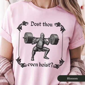 Pode incluir: Camiseta rosa com um gr&aacute;fico de um cavaleiro de armadura levantando pesos. O texto na camiseta diz "Dost thou even hoist?"