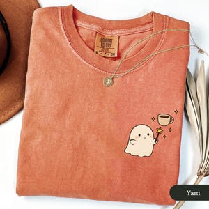 May include: A coral Comfort Colors t-shirt with a small graphic of a friendly ghost holding a magic wand next to a coffee cup. A gold necklace with a pendant rests on the shirt. The shirt is folded on a white surface.