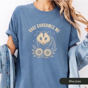 May include: Blue jean coloured t-shirt featuring the text "Rage Consumes Me" above an opossum graphic. The opossum is tan and grey, with cream-coloured flowers below. The t-shirt is a relaxed fit.