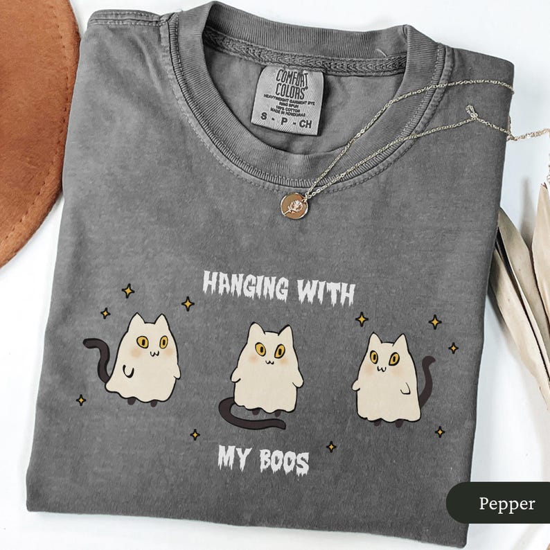 May include: A grey Comfort Colors t-shirt featuring the text "Hanging with My Boos" and a graphic of three ghost cats. A gold necklace with a pendant rests on the shirt. The shirt is size small.