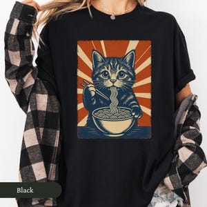 May include: Black t-shirt featuring a graphic of a cat eating ramen noodles with chopsticks. The cat is depicted in a vintage style with a red and beige background. The word "Black" is printed at the bottom.
