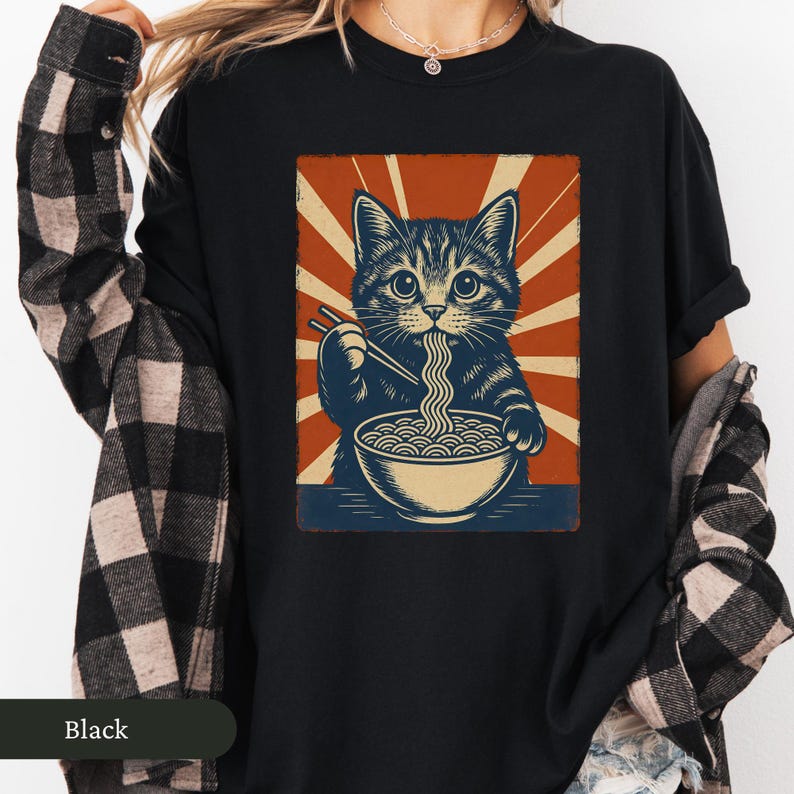 May include: Black t-shirt featuring a graphic of a cat eating ramen noodles with chopsticks. The cat is depicted in a vintage style with a red and beige background. The word "Black" is printed at the bottom.