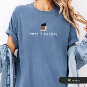 Może przedstawiać: Koszulka w kolorze niebieskiego jeansu z napisem "cats & books". Czarny kot siedzi na stosie książek.