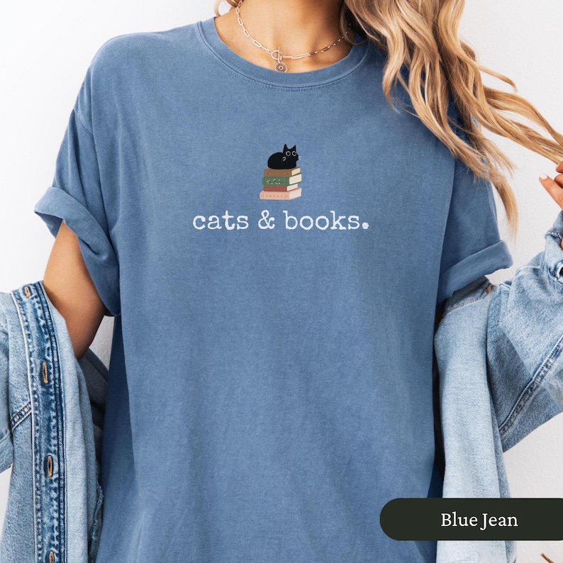 Może przedstawiać: Koszulka w kolorze niebieskiego jeansu z napisem "cats & books". Czarny kot siedzi na stosie książek.