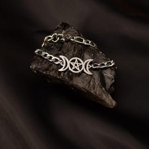 Könnte beinhalten: Ein silbernes Armband mit einem Design aus dreifachem Mond und Pentagramm. Das Armband liegt auf einem dunklen, strukturierten Stein vor schwarzem Hintergrund. Die Kettenglieder sind sichtbar.