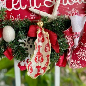 Può includere: Una ghirlanda natalizia festosa con un cartello di legno rosso che recita "Let it Snow" in lettere bianche. La ghirlanda è decorata con rami sempreverdi, nastri rossi e un ornamento a tema aragosta. Una stella marina e ornamenti a fiocco di neve aggiungono al tema delle vacanze.