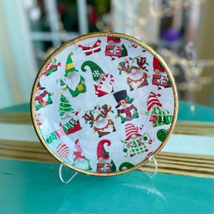 May include: A round decorative plate with a gold rim, featuring a pattern of Christmas gnomes in various poses. The gnomes wear red, green, and white outfits, some holding gifts or Christmas trees. The plate is displayed on a clear stand.