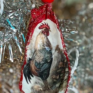 Puede incluir: Adorno navideño decorativo con una ilustración de un gallo. El adorno es una concha de ostra con un borde rojo brillante y una bola roja en la parte superior. El gallo está representado en tonos marrones, grises y blancos.