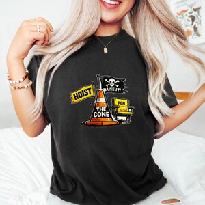 May include: Black t-shirt featuring a graphic with a traffic cone, a yellow chair, and flags that read "HOIST" and "RAISE IT!". The cone has the text "THE CONE". The shirt has a crew neck.