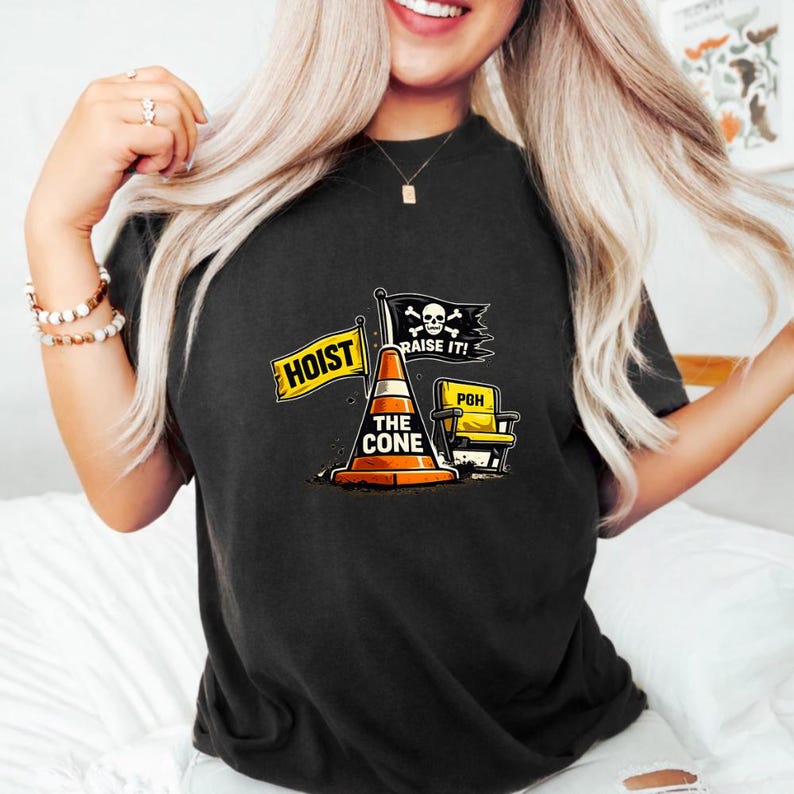 May include: Black t-shirt featuring a graphic with a traffic cone, a yellow chair, and flags that read "HOIST" and "RAISE IT!". The cone has the text "THE CONE". The shirt has a crew neck.