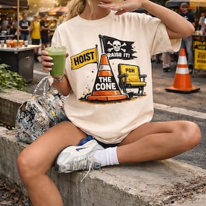 May include: Cream-colored t-shirt with a graphic featuring a traffic cone, a yellow chair, and the text "HOIST THE CONE" and "RAISE IT!". The image also includes a black flag with a skull and crossbones. The person is wearing a blue cap.