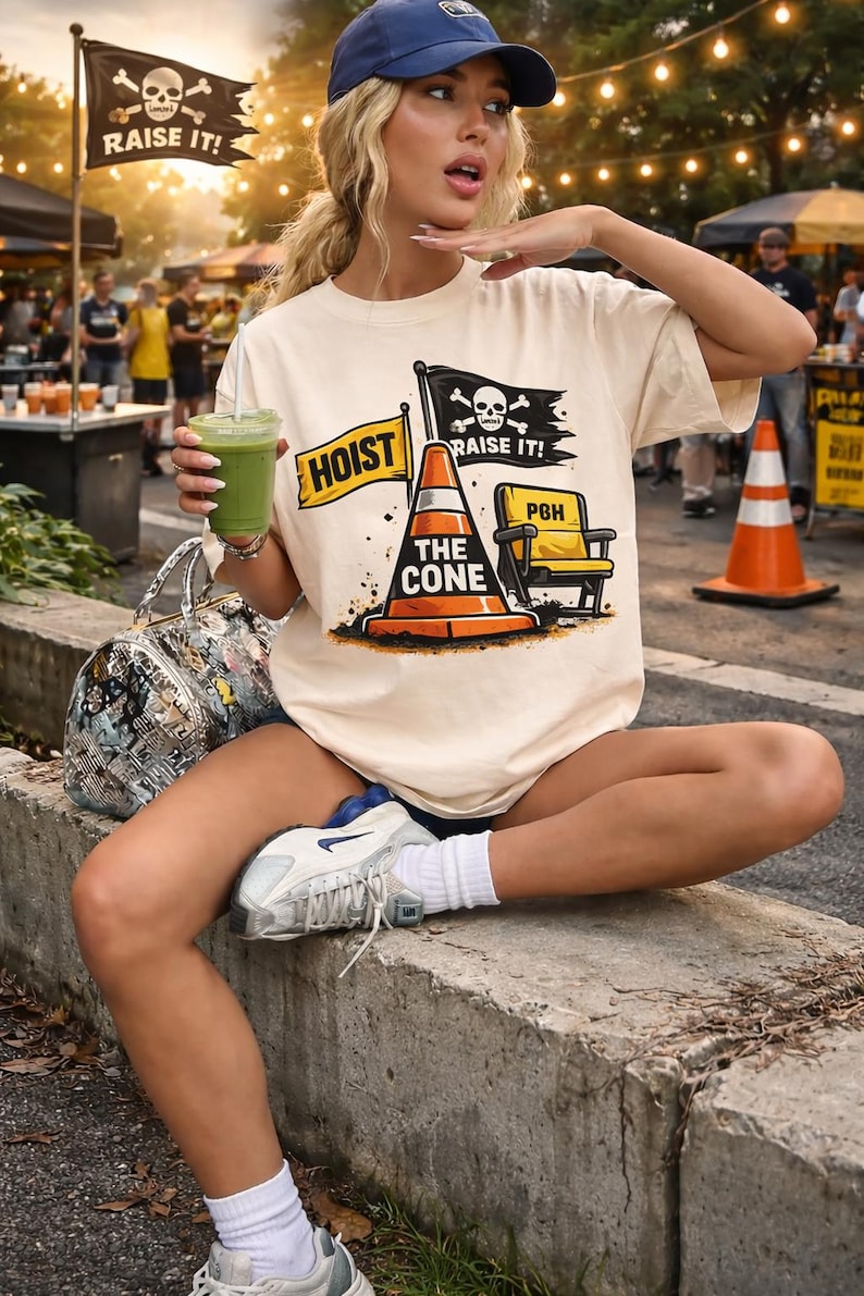 May include: Cream-colored t-shirt with a graphic featuring a traffic cone, a yellow chair, and the text "HOIST THE CONE" and "RAISE IT!". The image also includes a black flag with a skull and crossbones. The person is wearing a blue cap.