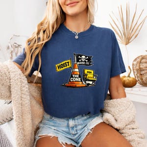 May include: Navy blue t-shirt featuring a humorous graphic. The design includes a traffic cone, a yellow chair, and flags with the text "HOIST" and "RAISE IT!". The shirt's design has a pirate theme with a skull and crossbones.