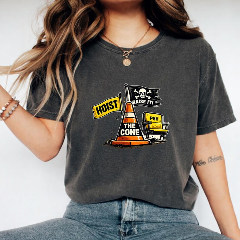 May include: Dark grey t-shirt featuring a graphic with a traffic cone, a yellow flag that says "HOIST", and a black flag with a skull and crossbones that says "RAISE IT!". The text "THE CONE" is on the cone.