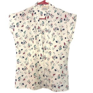 Peut inclure: Un chemisier sans manches de couleur crème avec un motif floral. Le motif présente des feuilles et des formes de fleurs rouges, bleues et vertes. Le chemisier est suspendu à un cintre rouge.