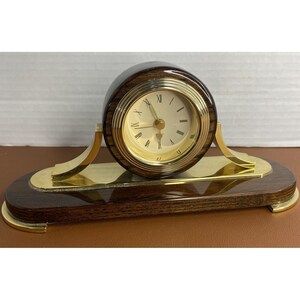 May include: A desk clock featuring a circular dark wood casing with a gold-toned bezel and a cream-colored face with Roman numerals. The clock is mounted on a polished gold-toned metal base attached to an oval dark wood plinth.