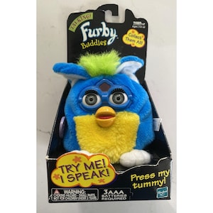May include: A blue and yellow Furby Buddies plush toy with green hair, large grey eyes, and a yellow beak, packaged in a black cardboard box with the text Talking! Furby Buddies, Try Me! I Speak!, and Press my tummy! visible on the front.