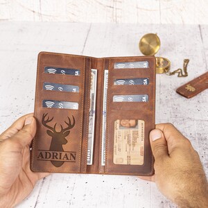 May include: Open brown leather wallet with multiple card slots, a clear ID window, and a cash compartment. The wallet features a deer head design and the name "ADRIAN" engraved on the front. The wallet is open, revealing cash and credit cards.