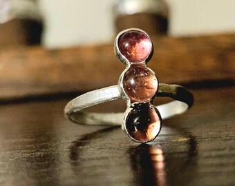 May include: A silver ring with three pink gemstones set in a row. The ring is on a wooden surface.