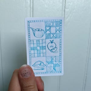 May include: A rectangular card with a blue and white quilt-like design. The card features hand-drawn illustrations of a teapot, apple slice, plate of food, and a grid pattern. The card is held by a person.