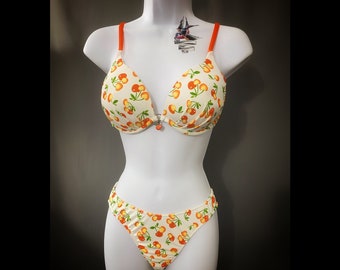 May include: White bikini with orange straps and a cherry print. The bikini top has a push-up style and the bottom is a cheeky cut.