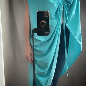 May include: A turquoise blue, loose-fitting, long tunic top with a gathered detail at the neckline. The top is worn with dark blue jeans.
