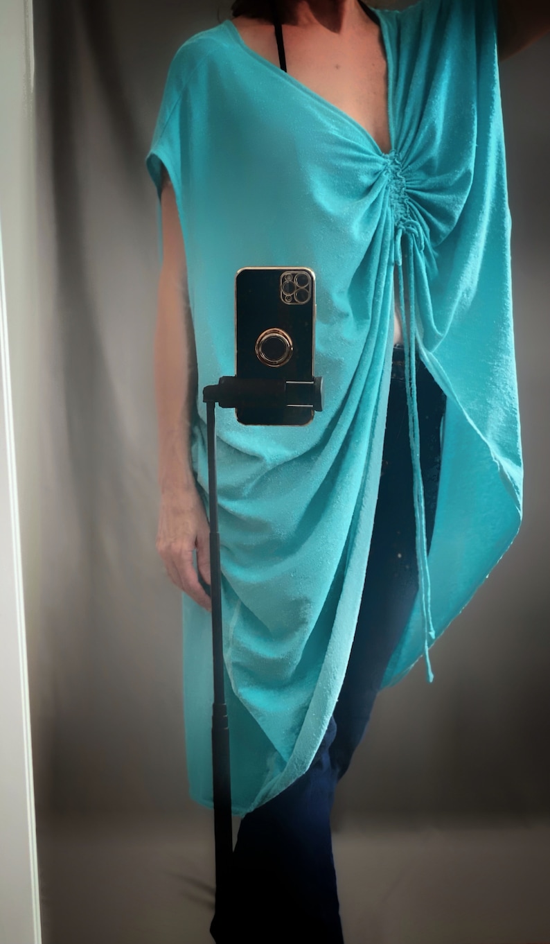May include: A turquoise blue, loose-fitting, long tunic top with a gathered detail at the neckline. The top is worn with dark blue jeans.