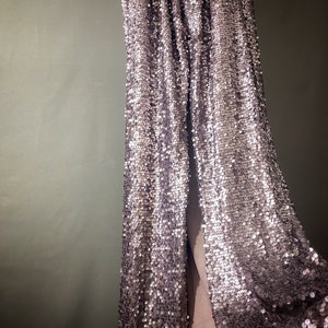 May include: A long, flowing skirt made of silver sequins. The skirt is draped over a surface and the sequins catch the light, creating a shimmering effect.