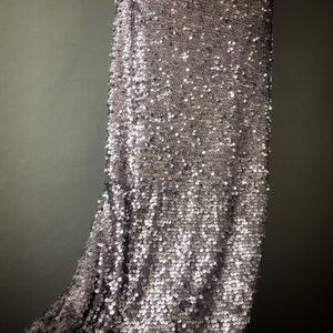 May include: A silver sequined fabric with a subtle sheen. The fabric is draped and shows the texture of the sequins.
